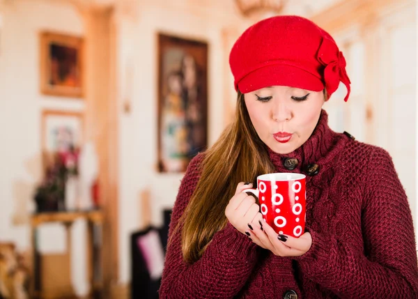 Bella giovane donna bruna che indossa maglione rosso e beenie, tenendo in mano una tazza di bevanda calda, soffiando sul liquido — Foto Stock