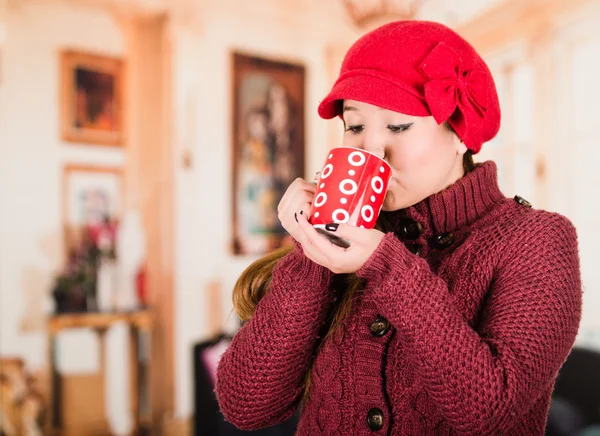 Muito jovem morena mulher vestindo camisola vermelha e beenie, tomando um gole de xícara de bebida quente — Fotografia de Stock