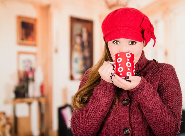 Muito jovem morena mulher vestindo camisola vermelha e beenie, tomando um gole de xícara de bebida quente — Fotografia de Stock