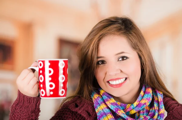 Mooie jonge brunette vrouw dragen rode jas en sjaal, houden kopje warme drank glimlachend naar camera — Stockfoto