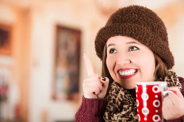 Bella giovane donna bruna che indossa giacca rossa, cappello e sciarpa, con in mano una tazza di bevanda calda sorridente — Foto Stock