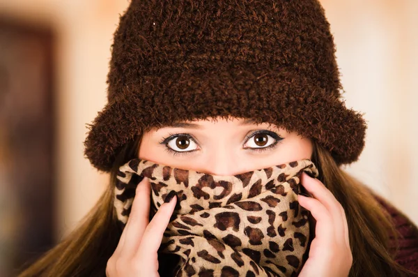 Pretty young brunette woman wearing brown hat and scarf covering up face revealing only eyes, staring straight into camera — Stock Photo, Image