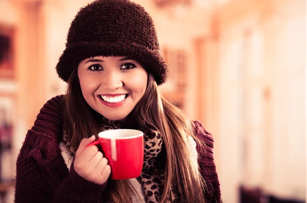 Krásná mladá brunetka, která nosí červený kabát, klobouk a šátek, držící hrnek horké nápoje s úsměvem na kameru — Stock fotografie