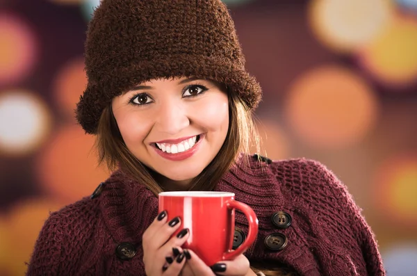 Pretty young brunette woman wearing red jacket, hat and scarf, holding cup of hot beverage smiling to camera