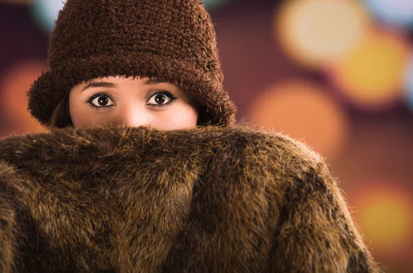 Bella giovane donna bruna in possesso di giacca stile pelliccia che copre metà faccia, cappello marrone in posa con sfocata luce gocce di sfondo — Foto Stock