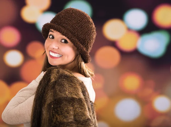 Pretty young brunette woman wearing fur style jacket, hat and scarf posing happily with a glamorous blurry light drops background — Stock Photo, Image