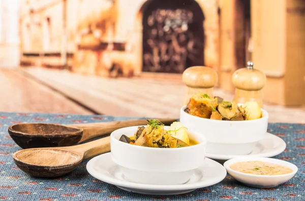 Serviço elegante de sopa de batata com carne e legumes, servido em tigelas brancas sentadas na mesa, toalha de mesa azul, saleiros pimenta, aconchegante restaurante fundo — Fotografia de Stock