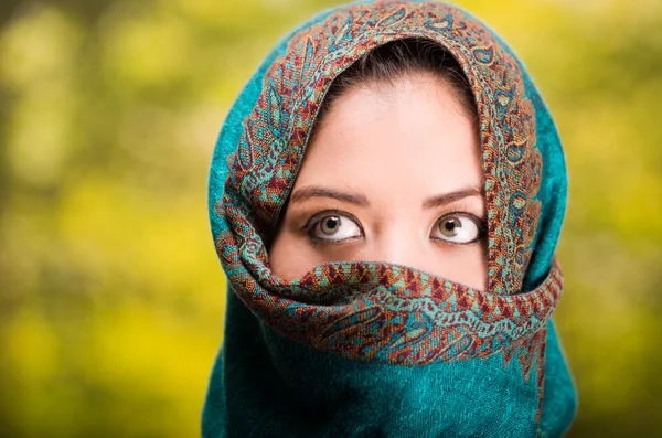 Mujer vistiendo azul, gris y marrón bufanda de color que cubre la cara solo revelando hermosos ojos verdes, fondo del jardín — Foto de Stock