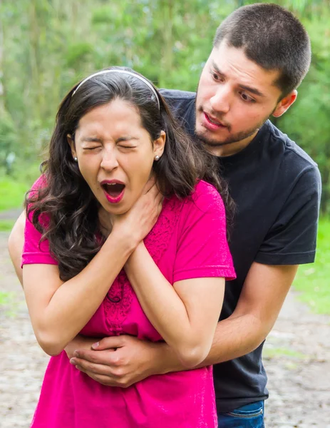 Young woman choking with man standing behind performing heimlich maneuver, park environment and casual clothes — Stock Photo, Image