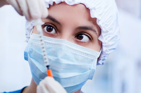 Closeup headshot nurse wearing bouffant cap and facial mask holding up syringe needle for camera