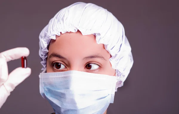 Nurse wearing bouffant cap and facial mask holding up brown pill capsule for camera, grey background — Stock Photo, Image