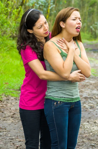 Mujer joven ahogándose con la señora de pie detrás de la realización de maniobra heimlich, el entorno del parque y la ropa casual —  Fotos de Stock