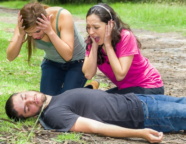 Hombre joven acostado con emergencia médica, dos mujeres jóvenes actuando histéricamente, ambiente al aire libre —  Fotos de Stock