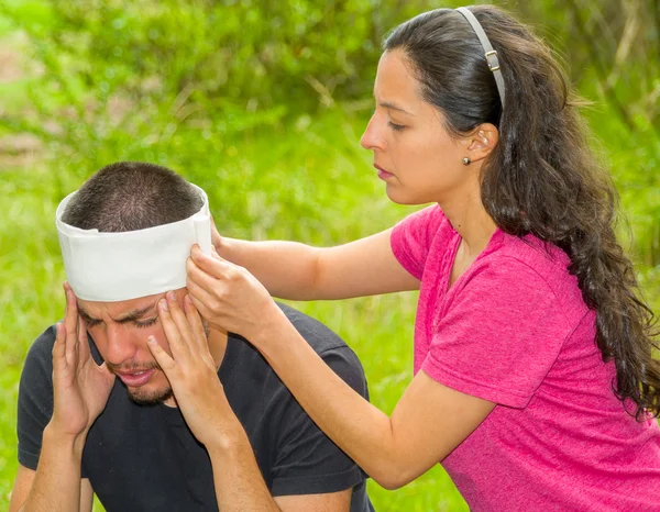 Hombre joven con lesión en la cabeza que recibe tratamiento y vendaje alrededor del cráneo de la mujer, ambiente al aire libre — Foto de Stock