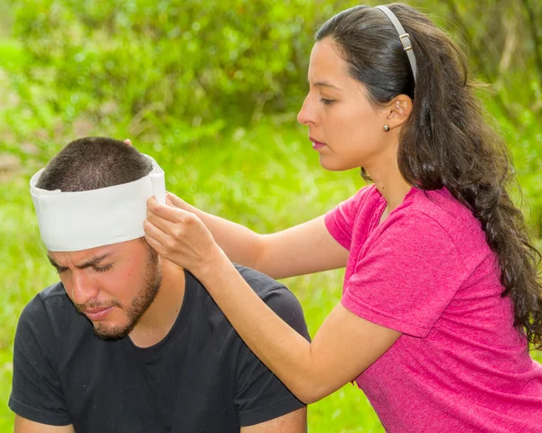 Hombre joven con lesión en la cabeza que recibe tratamiento y vendaje alrededor del cráneo de la mujer, ambiente al aire libre — Foto de Stock