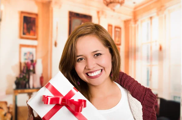 Brunette with beautiful smile holding up present, white wrapping and red ribbon, household background — Stock Photo, Image