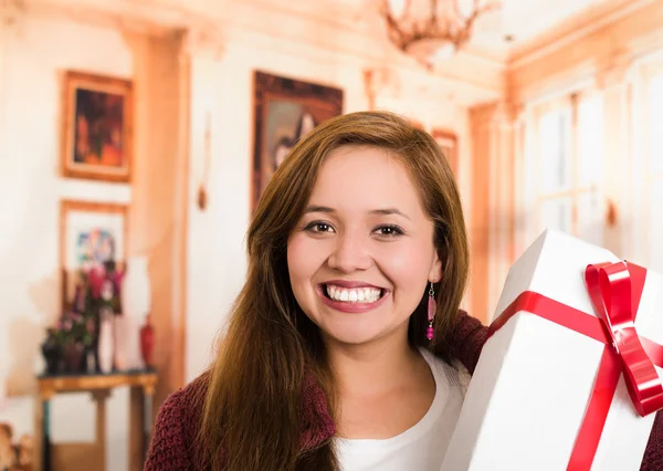 Brunette with beautiful smile holding up present, white wrapping and red ribbon, household background — Stock Photo, Image