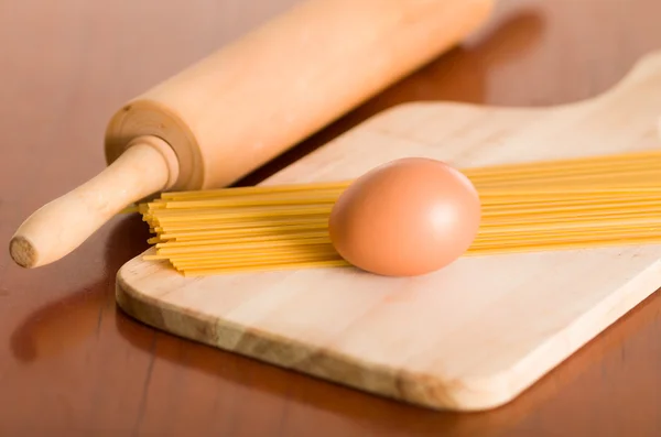 Wooden chopping board with egg and uncooked pasta piled up, rolling pin on desk next to it