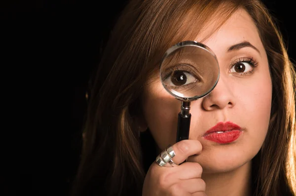 Young woman facing camera holding magnifying glass over right eye creating enhanced effect, black background — Stock Photo, Image