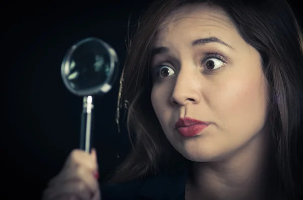 Young woman holding magnifying glass in front of eye and looking focused, black background — Stock Photo, Image