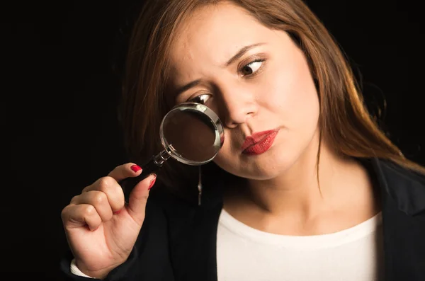 Young woman holding magnifying glass in front of face, slightly tilting head forward looking through it, black background — Stock Photo, Image
