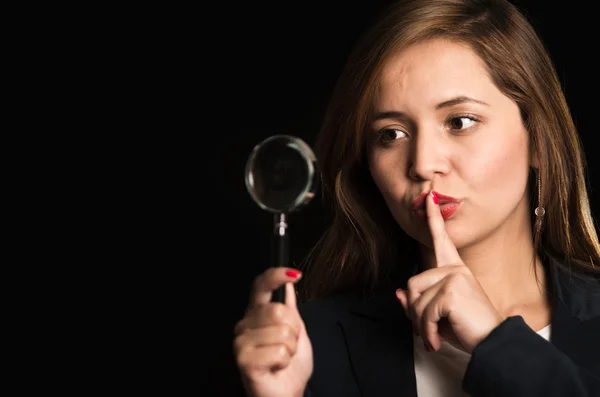 Young woman standing wearing casual clothes, holding magnifying glass with one hand, index finger across lips — Stock Photo, Image