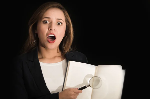 Young brunette woman facing camera holding up open book and magnifying glass above pages — Stock Photo, Image