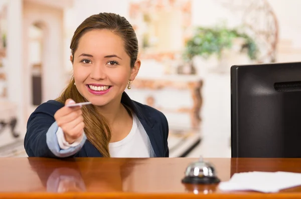 Unga brunett hotellreceptionist med vänliga leende lämnar över nyckeln till klienten över skrivbord, kunder synvinkel — Stockfoto