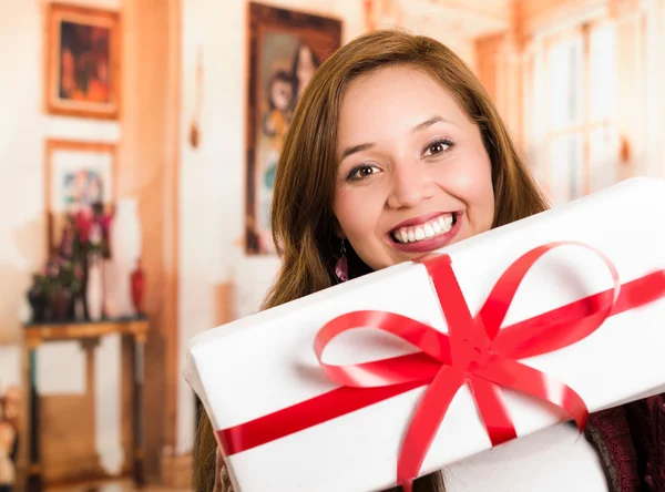 Brunette with beautiful smile holding up present happily posing for camera, white wrapping and red ribbon, household background — Stock Photo, Image