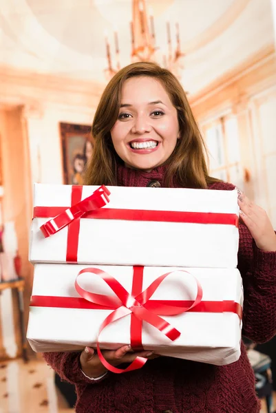 Morena com belo sorriso segurando presente feliz posando para câmera, embrulho branco e fita vermelha, fundo doméstico — Fotografia de Stock