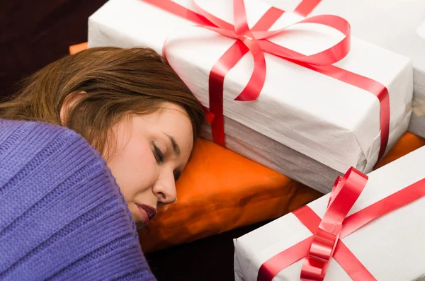 Morena cubierta con ropa morada durmiendo, cabeza descansada entre regalos, papel de regalo blanco y cinta roja — Foto de Stock