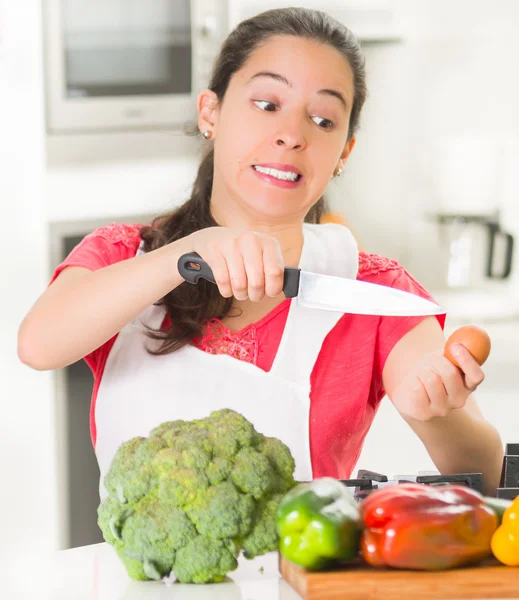 年轻的女厨师举起菜刀和鸡蛋，与面部表情，在前面的桌子上的蔬菜进行交互 — 图库照片