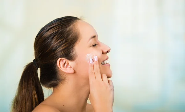 Bella giovane donna ispanica sana con le spalle nude, sorridente felicemente e applicare la crema per il viso con le mani — Foto Stock