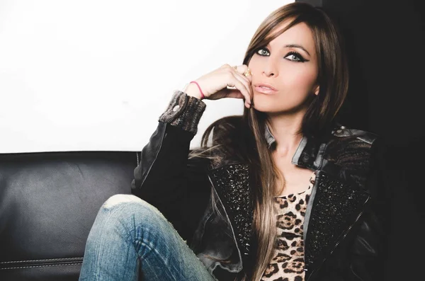 Sexy brunette wearing denim jeans, leopard top and leather jacket, sitting in sofa posing seductively for camera — Stock Photo, Image