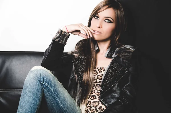 Sexy brunette wearing denim jeans, leopard top and leather jacket, sitting in sofa posing seductively for camera — Stock Photo, Image