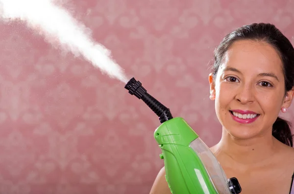 Brunette woman holding steam cleaner machine and vapor coming out, smiling to camera, pink background — Stock Photo, Image