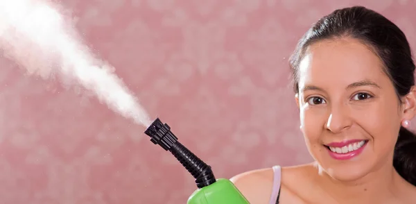 Brünette Frau hält Dampfreinigungsmaschine und Dampf, der herauskommt, lächelt in die Kamera, rosa Hintergrund — Stockfoto