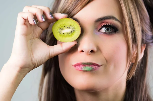 Headshot morena, olhar místico escuro e batom verde, cobrindo um olho com kiwi aberto, olhando para a câmera — Fotografia de Stock