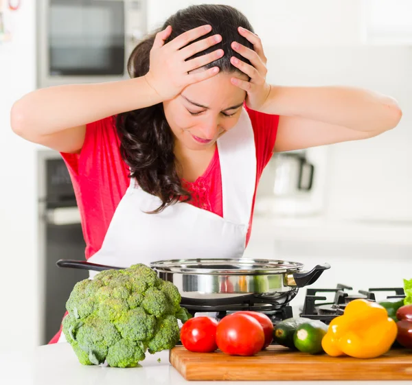 Giovane chef che tiene i capelli frustrati, espressione scoraggiata del viso, tavolo con bollitore e verdure — Foto Stock