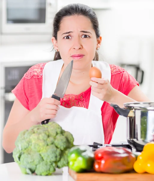 Jeune femme chef tenant couteau de cuisine et oeuf, interagissant avec des expressions faciales, légumes sur le bureau devant — Photo