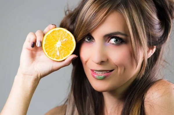 Headshot Brünette mit dunklem Mystique-Look und grünem Lippenstift, hält eine Orange neben das Gesicht und schaut in die Kamera, grauer Hintergrund — Stockfoto