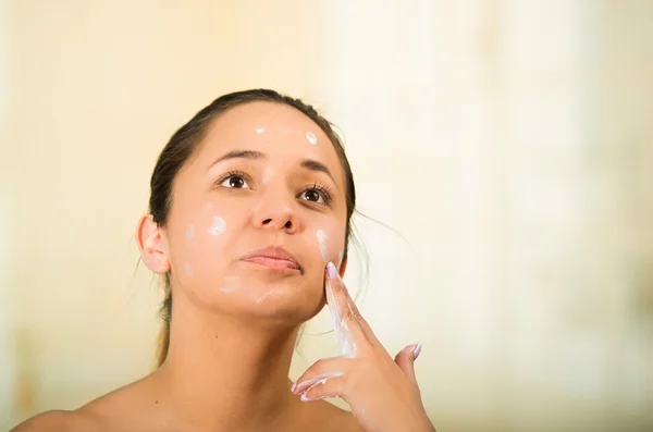 Pretty young healthy hispanic woman headshot with naked shoulders, applying cream to face using hands — Stock Photo, Image