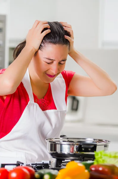 Jeune femme chef tenant les cheveux dans la frustration, expression faciale découragée, table avec bouilloire et légumes — Photo