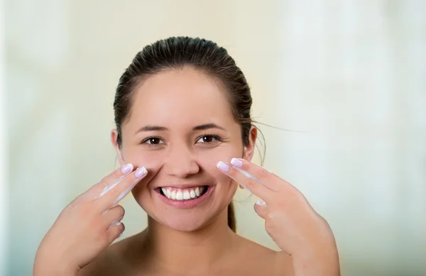 Pretty young healthy hispanic woman headshot with naked shoulders, smiling happily and applying cream to face using hands — Stock Photo, Image