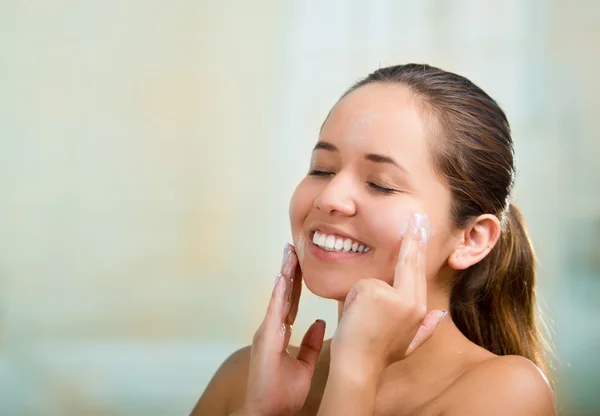 Pretty young healthy hispanic woman headshot with naked shoulders, smiling happily and applying cream to face using hands — Stock Photo, Image