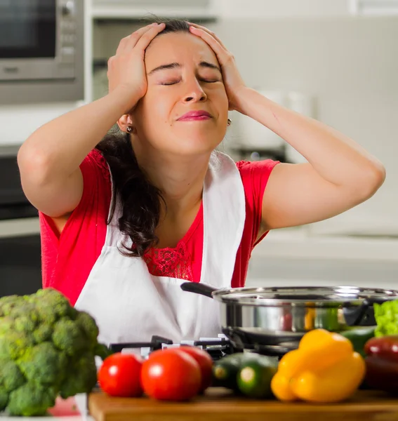 Jeune femme chef tenant les cheveux dans la frustration, expression faciale découragée, table avec bouilloire et légumes — Photo