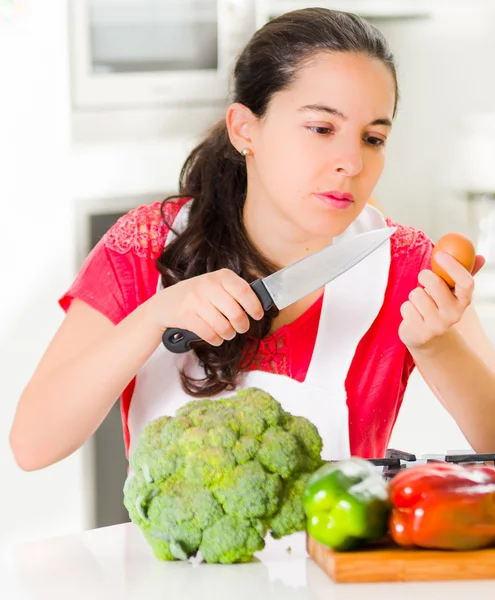 Young woman chef holding up kitchen knife and egg, interacting with facial expressions, vegetables on desk in front — Stock Photo, Image