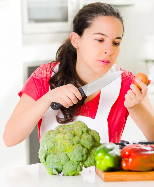 顔の表情、前の机の上の野菜と対話する包丁と卵を持って若い女性シェフ — ストック写真