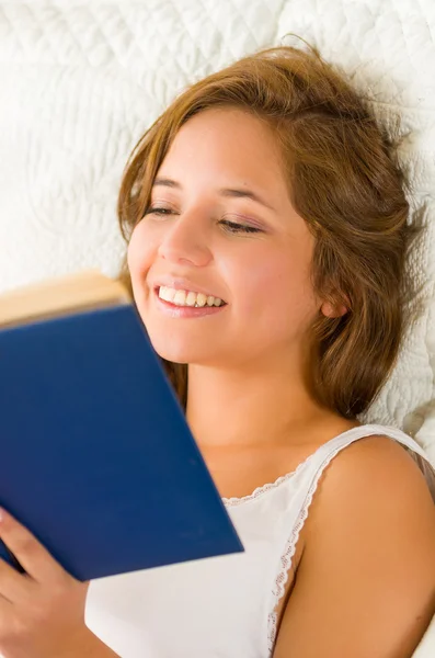 Jovem morena headshot na cama deitada confortavelmente cabeça descansando no travesseiro, lendo livro azul feliz — Fotografia de Stock