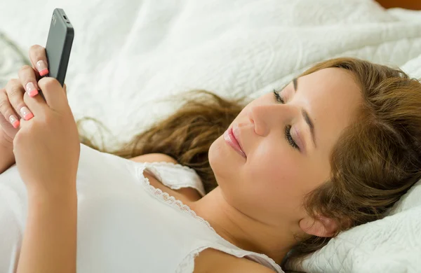 Mujer morena joven en la cama, descansando cómodamente sobre sábanas blancas y almohada, sosteniendo el teléfono móvil sonriendo — Foto de Stock